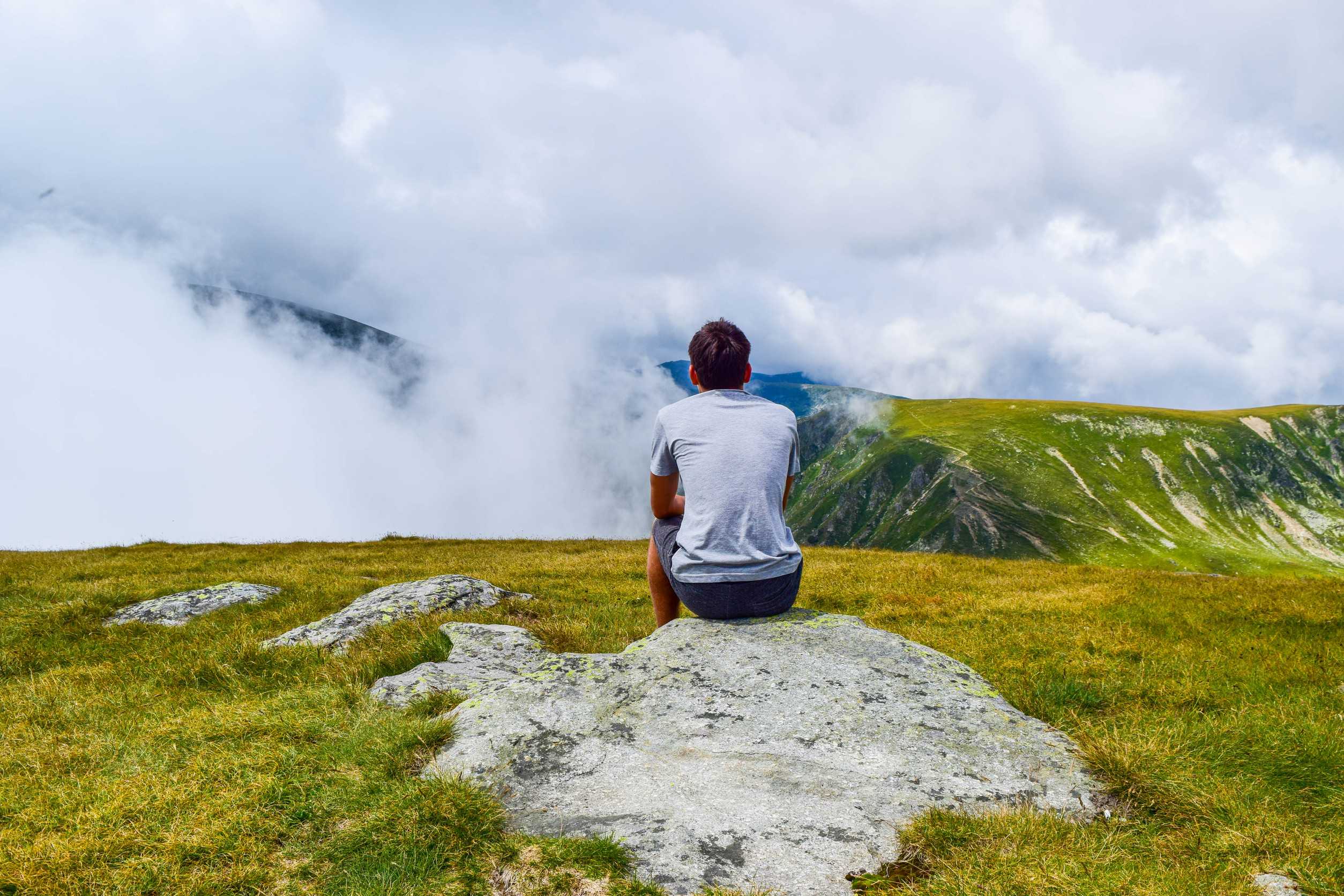 Guy Sits On A Stone Surrounded By Fog And Low Clouds Near Transalpina Road