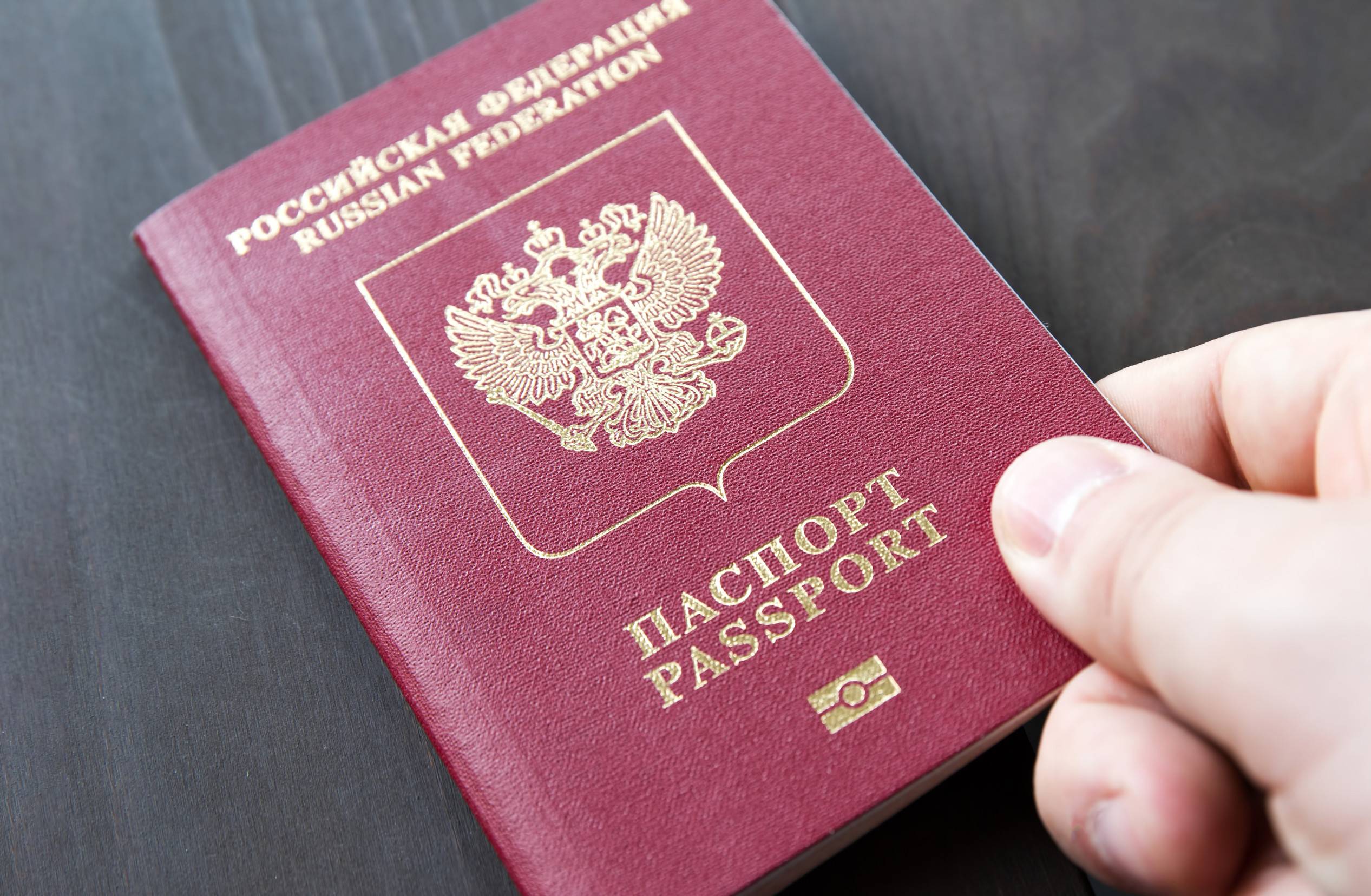 Male Hand On Top Of Red Passport On A Grey Wood Background